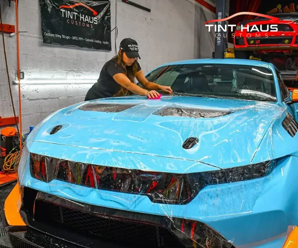 A professional technician applies a clear paint protection film to the hood of a bright blue sports car in an automotive shop environment.