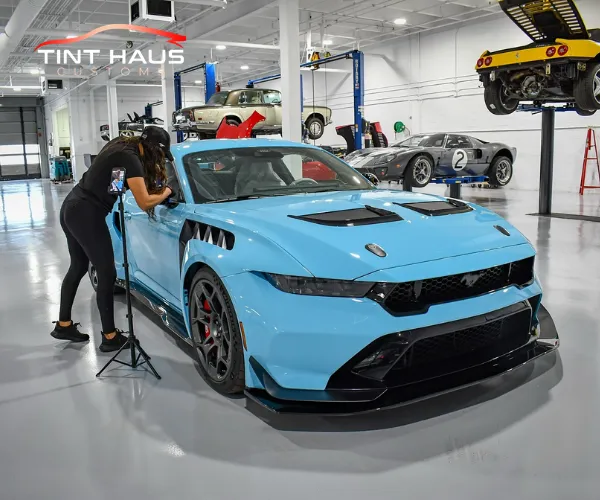 A technician works on the side window of a light blue performance vehicle in a spacious garage featuring multiple luxury cars on lifts.