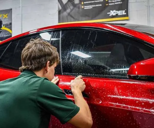 applying window tinting film to the side window of a red Tesla sports car inside a professional auto styling shop