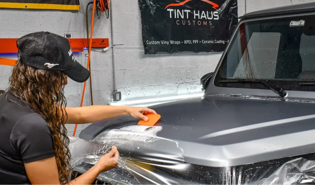 Installer using an orange squeegee to apply paint protection film on a vehicle hood.
