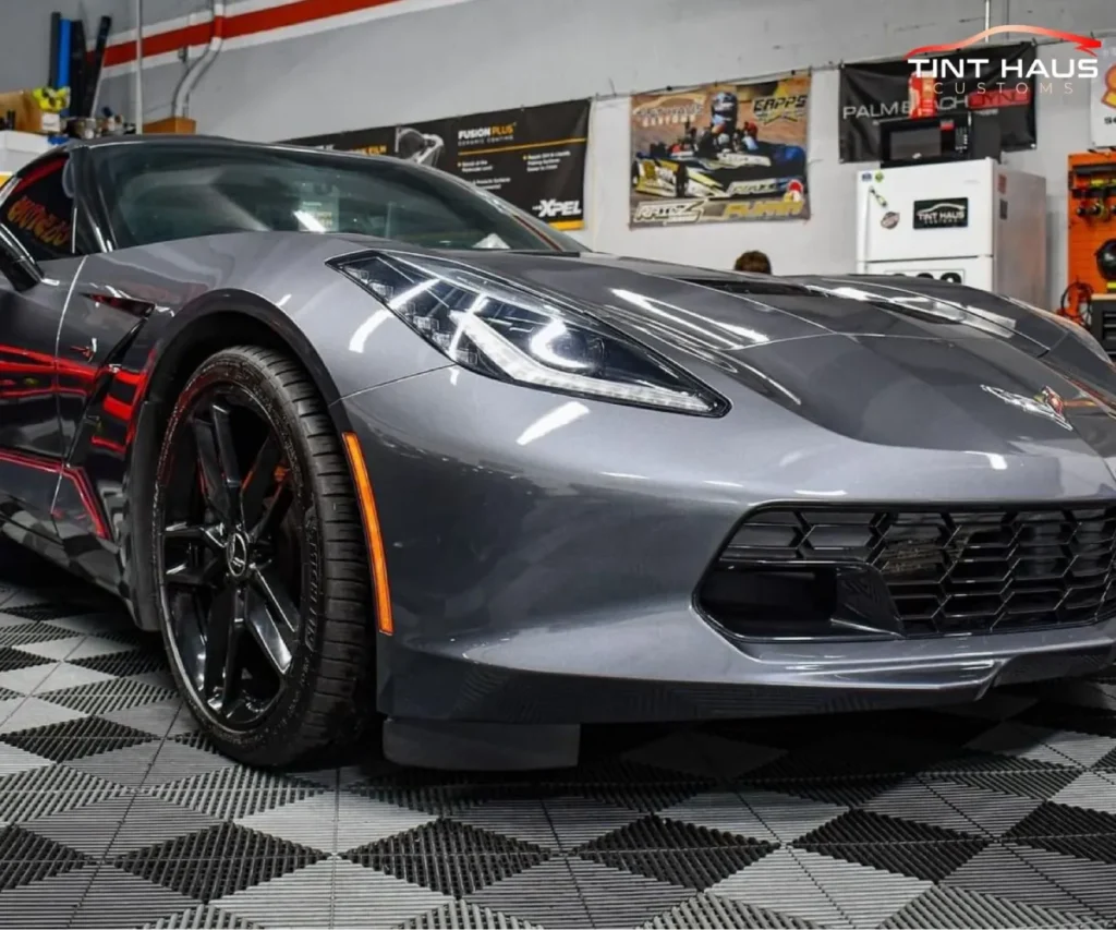 Low-angle close-up of the front headlight, black grille, and front wheel of a dark grey Chevrolet Corvette.